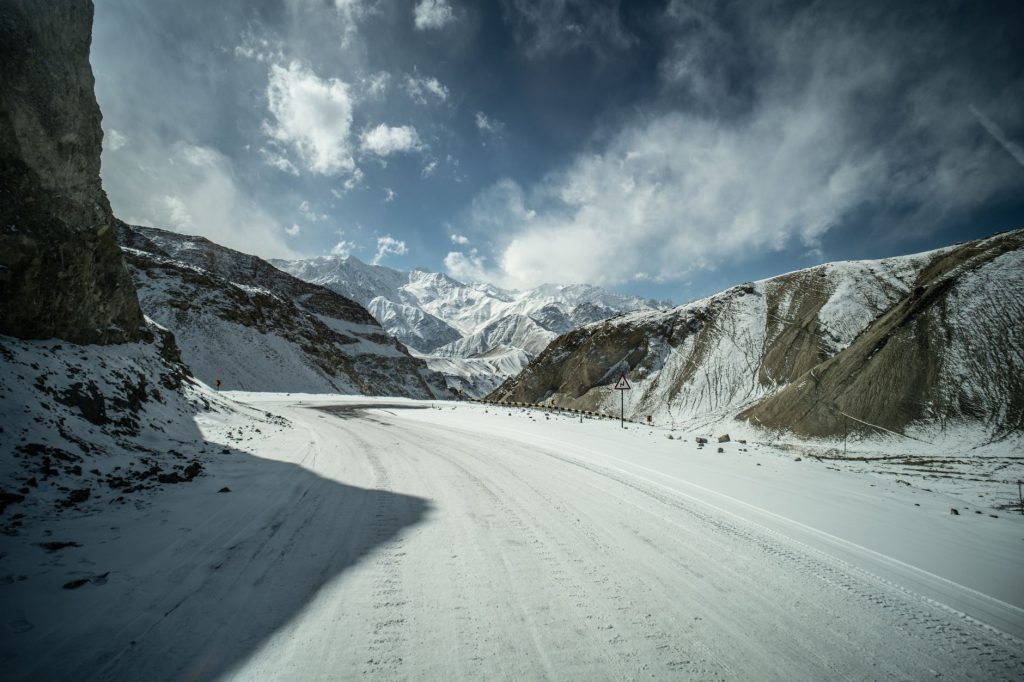 Winter scene of a snowy mountain road winding through rugged hills under a cloudy sky.