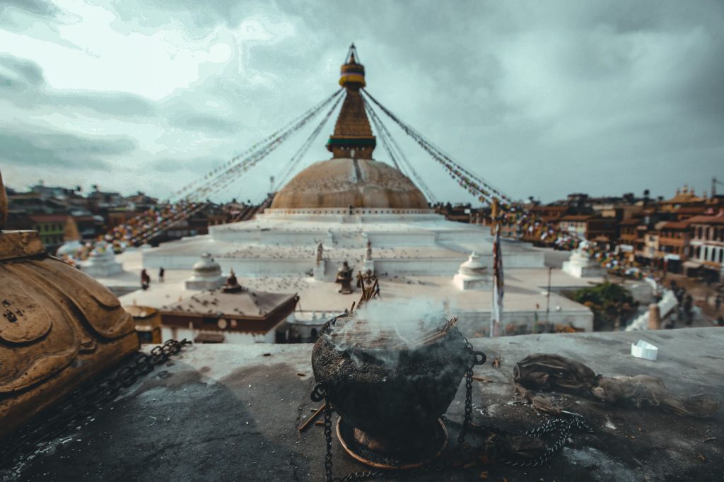 Incense burns before the iconic Boudhanath Stupa in Kathmandu, Nepal, under a cloudy sky.