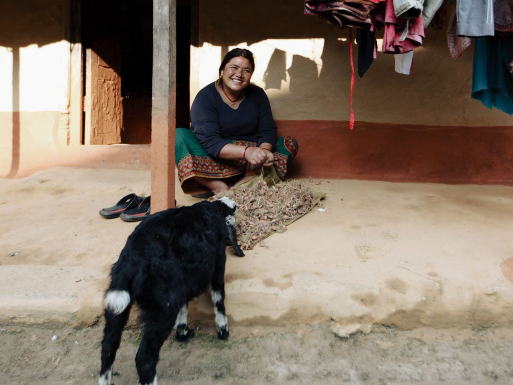 Smiling woman feeds a goat outside her home, laundry hanging above.