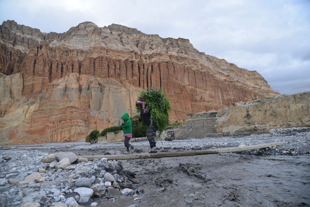 A mother and child walk across a rocky landscape in Upper Mustang, Nepal with a steep cliff backdrop.