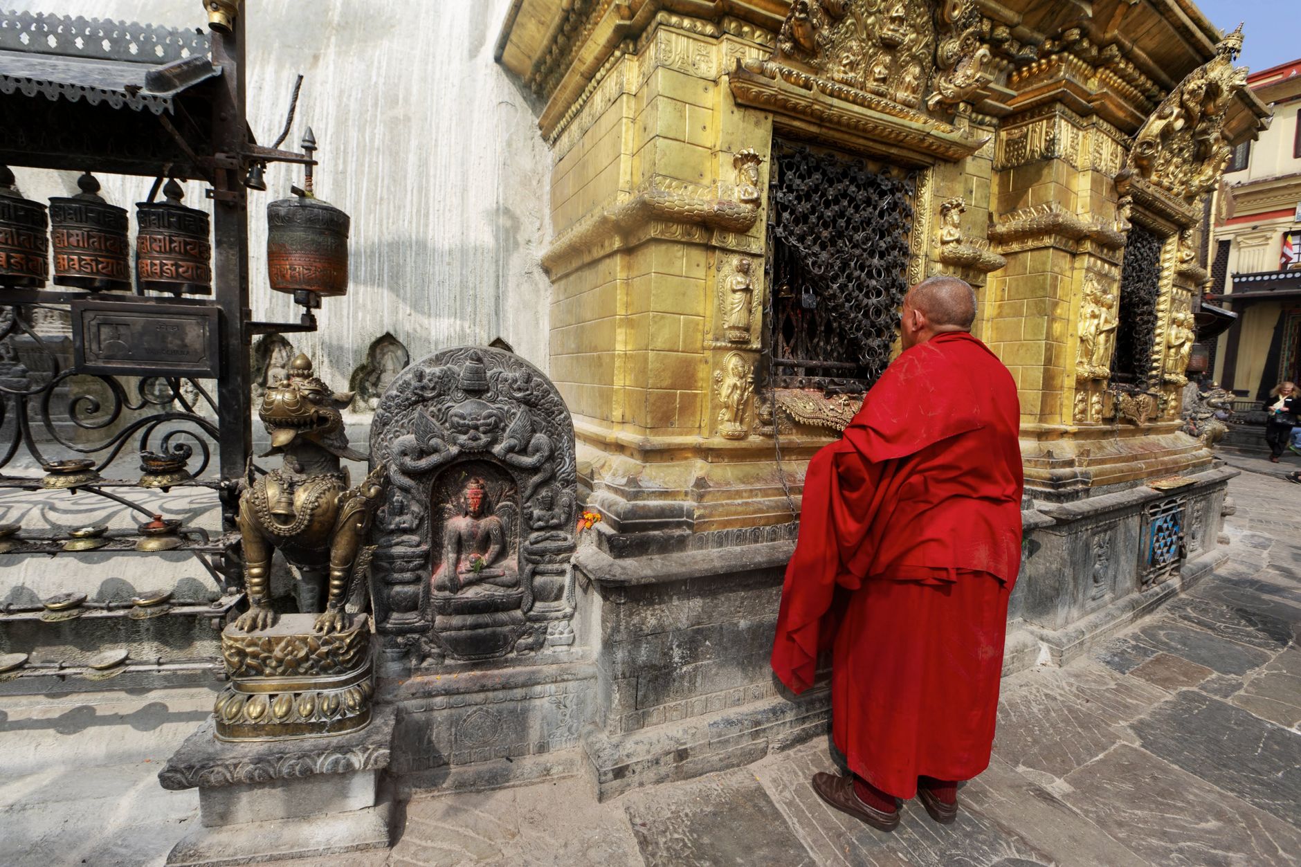 A Buddhist monk in vivid red robes visits a historic golden temple adorned with religious sculptures.