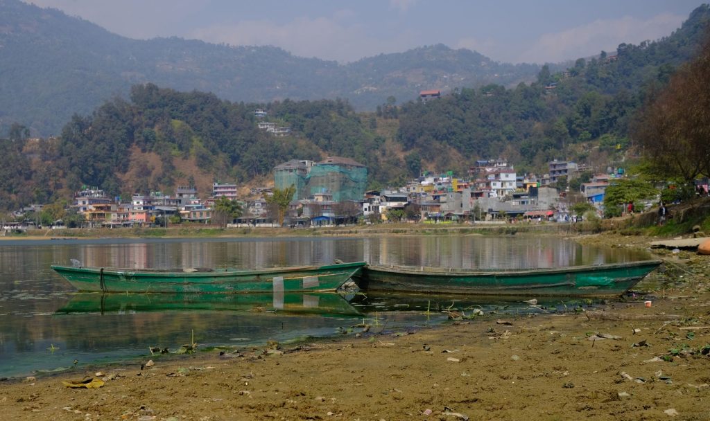 Green boats rest on Phewa Lake with Pokhara cityscape and hills in the background.
